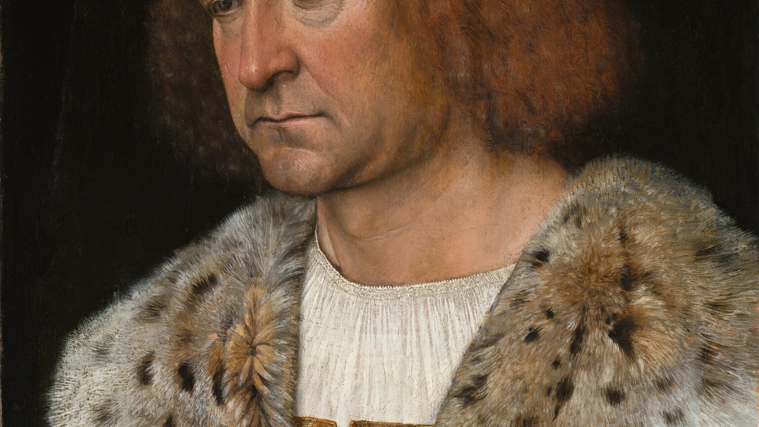 Shown from the chest up, a clean-shaven, pale-skinned man holds one hand to his chest and rests the other on a rug-covered ledge with the other in this vertical portrait painting. The man’s body is angled to our left, and he looks down in that direction with dark eyes under thick brows. Crow’s feet crinkle at the corners of his eyes, and there are dark hollows underneath. Soft jowls hang to create a slight double chin. His thin lips are set in a line over a wide, round chin, which is darkened with a five o’clock shadow. He has chin-length, reddish-blond hair, and straight, wispy bangs sweep loosely across his high forehead. He wears a white chemise under a gold, brocade garment, which is mostly hidden by a slate-blue cloak. The cloak is lined with spotted fur, which is turned over to create a wide collar across his shoulders. The sleeve on his left arm, to our right and closer to us, splits over a dusky-pink sleeve. Two gold buttons on the cloak there are decorated with thistle plants and blossoms. The fingers of his right hand, to our left, are gathered like a closed rose bud at his chest. He wears a gold ring on the index finger of the other hand, which rests on the end of the gray stone ledge so his fingertips brush the tapestry draped there. The man is lit from our left against a black background.