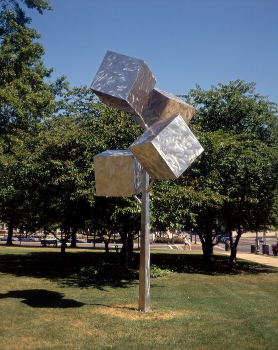 Four gleaming silver metal cubes are affixed to the top third of a tall pole with short arms like abstracted blossoms or tree limbs in this freestanding sculpture. The pole is planted in a grassy lawn and stands as tall as mature trees behind it. The smooth surface of the stainless steel appears streaked.