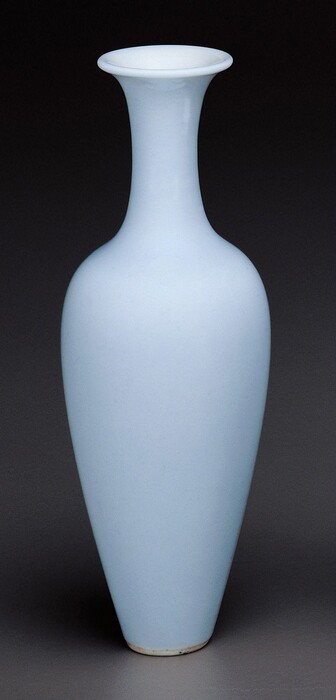 The image displays a single large vase in a pale blue color against a dark background. The vase has a slightly flared mouth, a rounded body, and tapers towards the base.