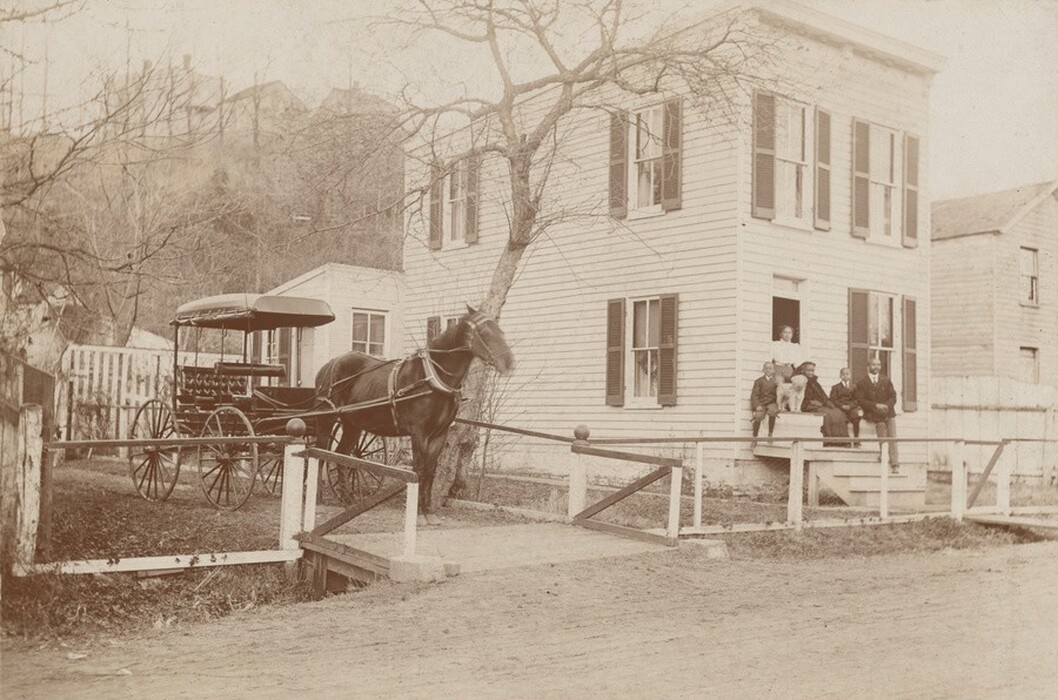 This is a photograph of a horse-drawn carriage near a two-story wooden house. The image captures a classic rural scene featuring a house with large windows framed by shutters and a simple gabled roof. The structure is positioned close to a dirt road, with a wooden fence lining the path leading to a small gate opening towards the home's entrance. A horse-drawn carriage, with its wheels and covered seats, is stationed near the house, tethered to a tree between the house and the foreground. In the background, there are bare trees and additional houses that provide depth and a rustic setting. The overall tone of the photograph suggests it's from an earlier time period, characterized by the sepia effect. Various details such as window details and the horse's harness are visible, offering insights into the period's lifestyle and architectural styles.