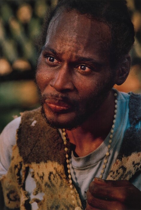 The image shows a person's upper body, focusing on the face and torso. The person is turned to the side with their head tilted up and eyes looking in the same direction. They have dark skin, expressive eyes, a trimmed beard, and dark hair pulled back. They wear a patterned garment, a light-colored shirt, and have beads around their neck, holding onto them with one hand. The background is softly blurred with a warm, natural ambiance.