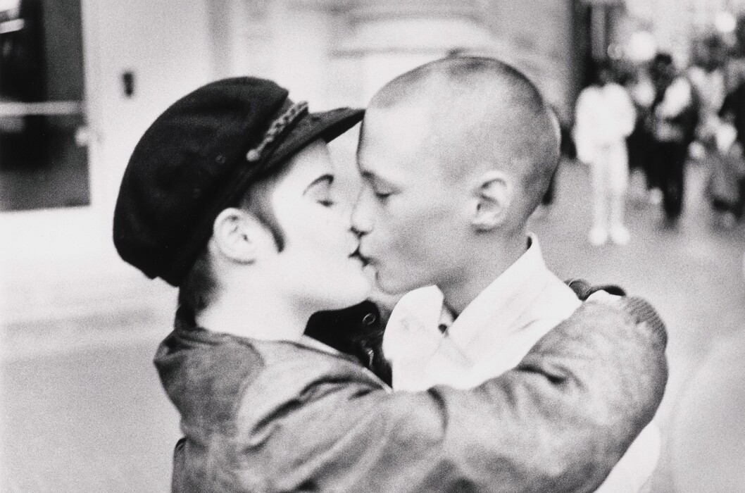 The image depicts two individuals in a close embrace, with their faces pressed together as they kiss. Only the upper half of their bodies is visible. The person on the left has short hair peeking out from under a cap with a decorative band. Their eyes are closed. The person on the right has very short hair and is wearing a white shirt with their eyes also closed. The background is blurred, suggesting a street scene with indistinct figures in the distance. The focus remains on the two people in the foreground, while the background shows a busy or populated environment.