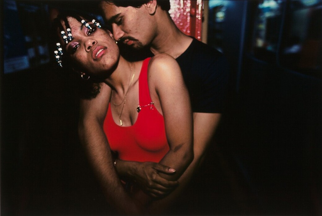 The image depicts two people embracing closely. The woman is in front, leaning back, while the man stands behind her, nuzzling her neck. The woman has short braided hair with white bead embellishments and is wearing a red top with a gold necklace. The man has short dark hair and facial hair, wearing a dark shirt. The background is dimly lit with blurred blue and red lights and some indistinct shapes.