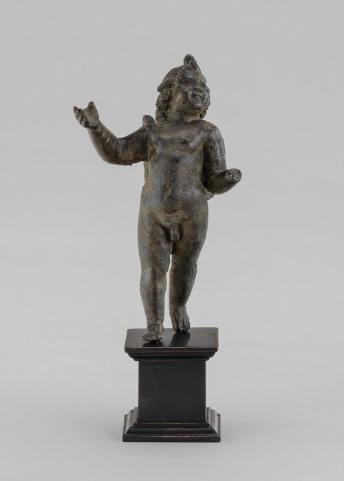 A smiling child stands facing us with arms lifted in this cast bronze, free-standing sculpture. The surface is rough and pitted throughout and oxidized to green in some areas. In this photograph, the boy’s body faces us but his head tips back, and he looks up and to the right. His right hand, to our left, is raised, palm up, to about shoulder height, and the other hand, held slightly lower, is missing. With rounded feet, the child stands on a pedestal base rubbed gold across moldings along the top and bottom.