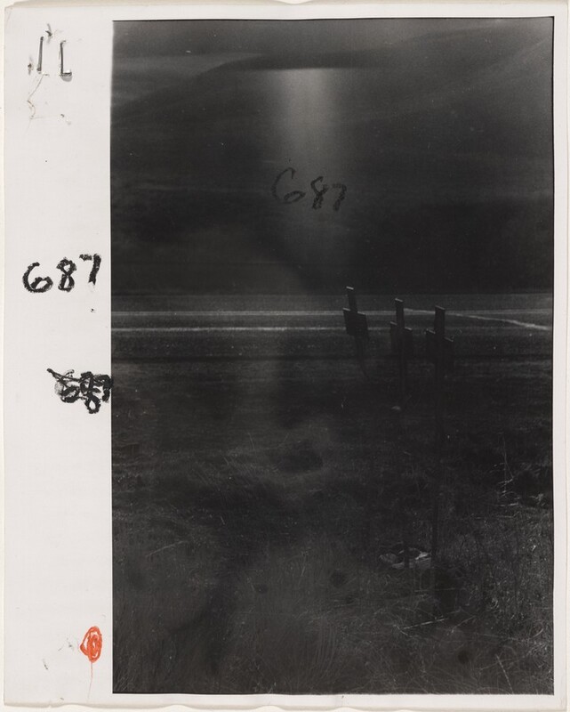 The image shows a monochrome view taken from a low perspective. The horizon is high in the image with a line of shadows or distant hills. Three objects resembling crosses or signposts are silhouetted against the background, slightly to the right. The color palette is dominated by shades of gray and black, creating a moody atmosphere. The photograph's textures and shadows create a visual impact. Handwritten scribbles and numbers are visible on the left and center of the image.