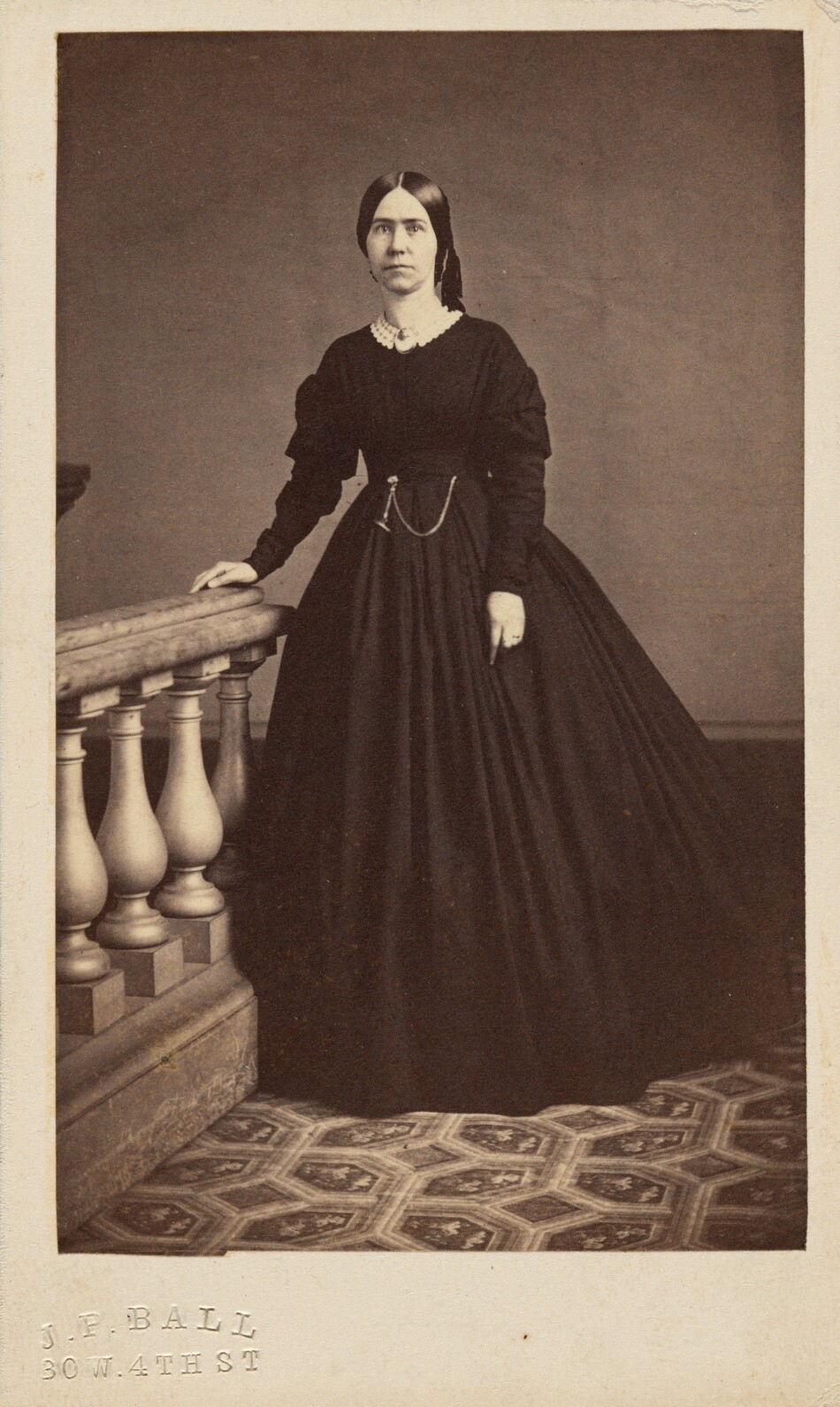 The image depicts a woman standing next to a wooden balustrade. She faces forward with her right hand resting on the railing. The woman has straight dark hair parted in the middle, and wears a full-skirted dress with a light-colored lace collar. She has a chain around her waist and a brooch at her neckline. The background is plain, with a patterned carpet on the floor. The photograph is vintage, with the name and address "J.P. Ball, 30 W. 4th St" stamped at the bottom.