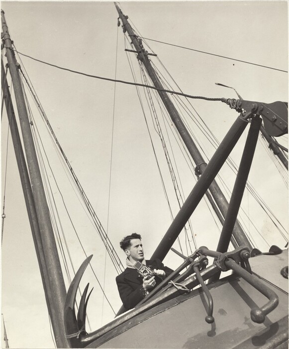 A man is on the deck of a ship that angles sharply to the left in this vertical black and white photograph. We look up at the man, who holds a boxy camera in both hands and looks off to our right almost in profile. His short, dark hair ruffles in the wind, and he wears a light-colored scarf and a dark coat. The ship’s anchor is slung over the side of the hull close to us, and two masts lunge vertiginously up toward the top left corner of the photograph against a clear sky.