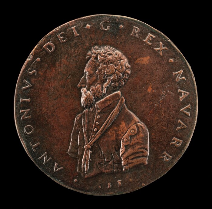 The sculpture depicted on the coin shows a profile portrait of a bearded man facing to the left. The man appears to be a high-ranking figure, as suggested by the detailed depiction of his clothes, which include a high-collared shirt with buttons and a decorative sash tied around the neck. The coin is made of bronze, giving it a coppery-brown color. The surface of the coin includes inscriptions along the edge, likely bearing the title and name of the figure, with text in Latin that includes "ANTONIVS" and "REX." The man's facial features are detailed, from his curly hair and prominent nose to his neatly trimmed beard. The relief of the coin gives a three-dimensional effect, making the figure stand out against the background.