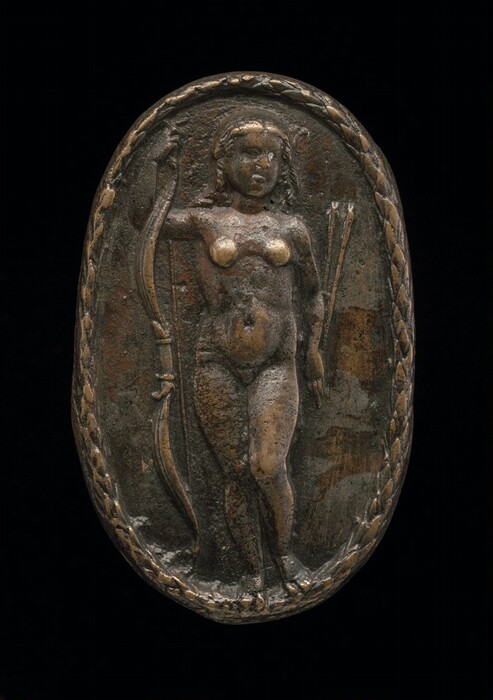 The sculpture depicts a human figure standing in a frontal pose with legs slightly apart, holding a bow in the left hand and a quiver of arrows in the right. The figure is nude and has distinct feminine features, such as breasts and flowing hair. The sculpture is crafted in a relief style within an oval frame with a braided or rope-like pattern along its edge. The material appears to be a type of metal with a worn, aged patina, displaying a base color of dark bronze or brownish-gold with areas where the metal’s natural color or polishing highlights shine through. The figure is rendered in a rough and textured manner, giving it an ancient and rustic appearance.