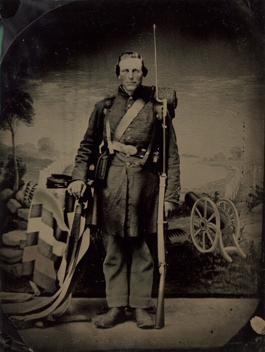 The image is a full-body portrait of a man standing upright. He is posed facing forward with a rifle in his right hand and his left hand resting on a folded flag draped over a nearby support. His facial features include a serious expression, high cheekbones, and closely cut hair. The man is dressed in a dark military uniform consisting of a buttoned coat, chest straps, and a belt at the waist. He wears dark trousers, sturdy leather boots, and has a backpack or bedroll strapped across his back. The background features a painted backdrop of a stylized landscape with foliage and a cannon to his side.