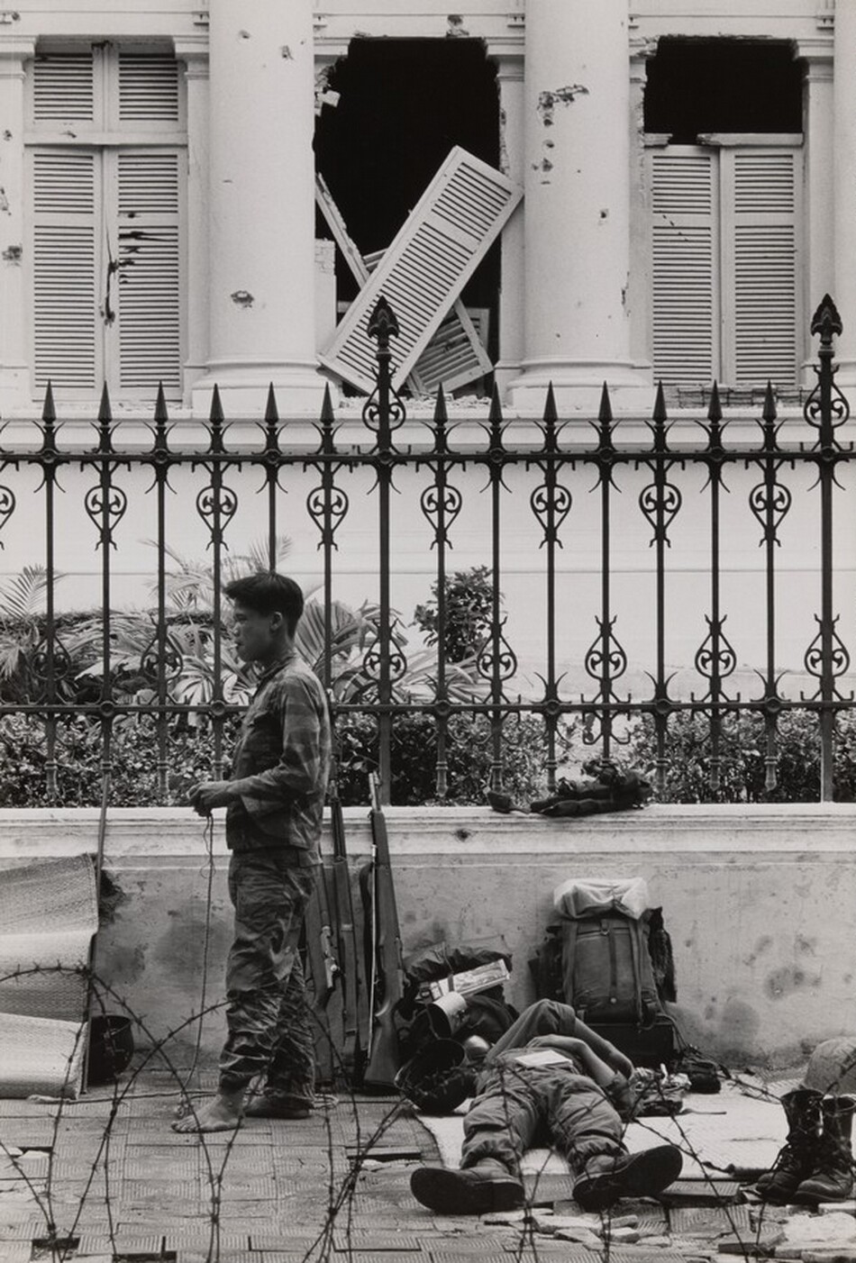 This grayscale photograph shows two people, perhaps soldiers, in front of a wrought-iron fence. One soldier stands upright, facing the left, dressed in dark clothing and holding a wire or cable, with three rifles propped against the wall beside him. Another soldier lies on the ground to the right, lying on his back with arms crossed, surrounded by military gear including a backpack and a pair of boots. Behind them, a large building has damaged shutters and a broken window, revealing a white facade with large columns. Coiled barbed wire is on the pavement closest to us.