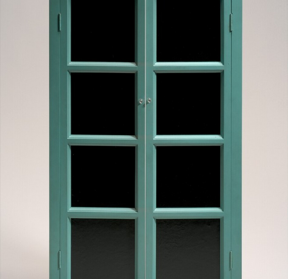 This free-standing sculpture is a French window with a teal-colored frame and black, leather-lined glass panes standing on a flat, shallow base. Each door is made up of four, vertically stacked, equally sized panes. The doors are hinged on a narrow frame and open with small, clear knobs placed next to the second pane down from the top. The doors are slightly ajar. Black writing in capital block letters on the top surface of the base is legible in this photograph. It reads, “FRESH WIDOW COPYRIGHT ROSE SELAVY 1920.”