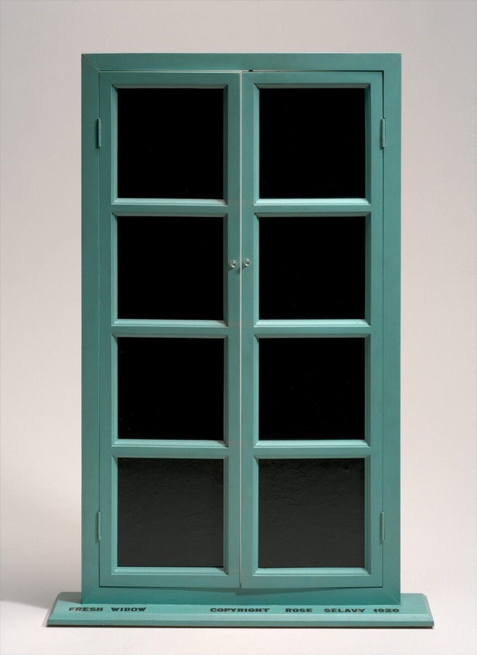 This free-standing sculpture is a French window with a teal-colored frame and black, leather-lined glass panes standing on a flat, shallow base. Each door is made up of four, vertically stacked, equally sized panes. The doors are hinged on a narrow frame and open with small, clear knobs placed next to the second pane down from the top. The doors are slightly ajar. Black writing in capital block letters on the top surface of the base is legible in this photograph. It reads, “FRESH WIDOW COPYRIGHT ROSE SELAVY 1920.”