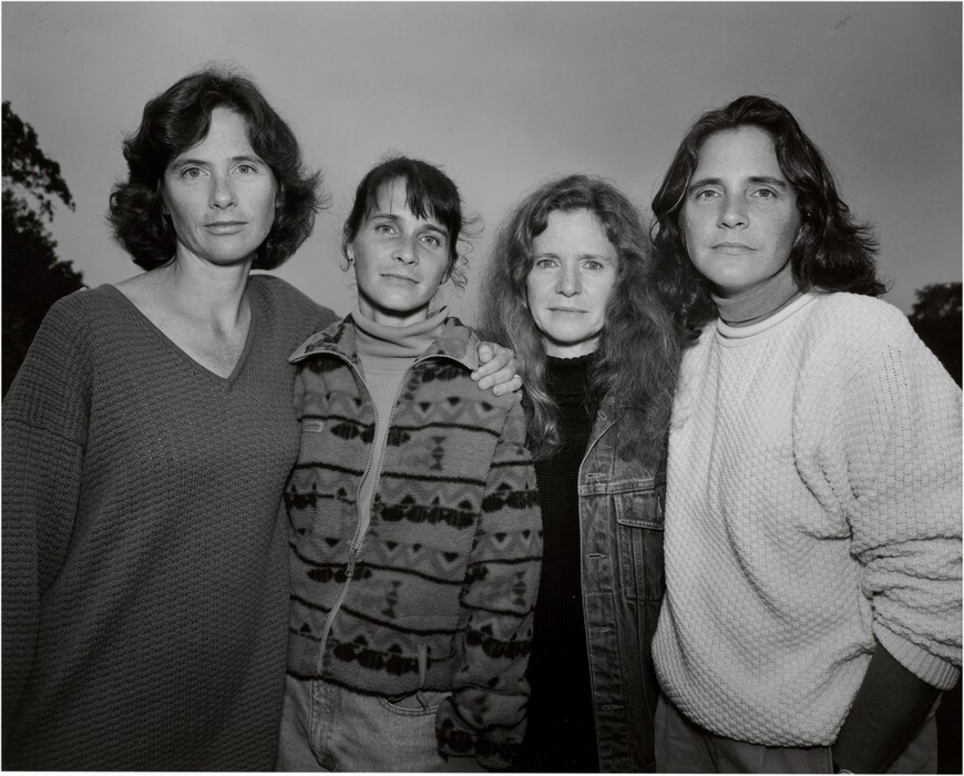 Four women are standing close together in a line. The woman on the left has medium-length wavy hair and is wearing a loose-knit sweater. The second woman has hair pulled back with bangs and is wearing a patterned jacket over a turtleneck. The third woman has long, curly hair and wears a denim jacket over a dark top. The woman on the right has shoulder-length wavy hair and is wearing a textured light sweater over a top with a dark collar. The backdrop is a natural environment, likely outdoors, with tree tops visible under a cloudy sky.