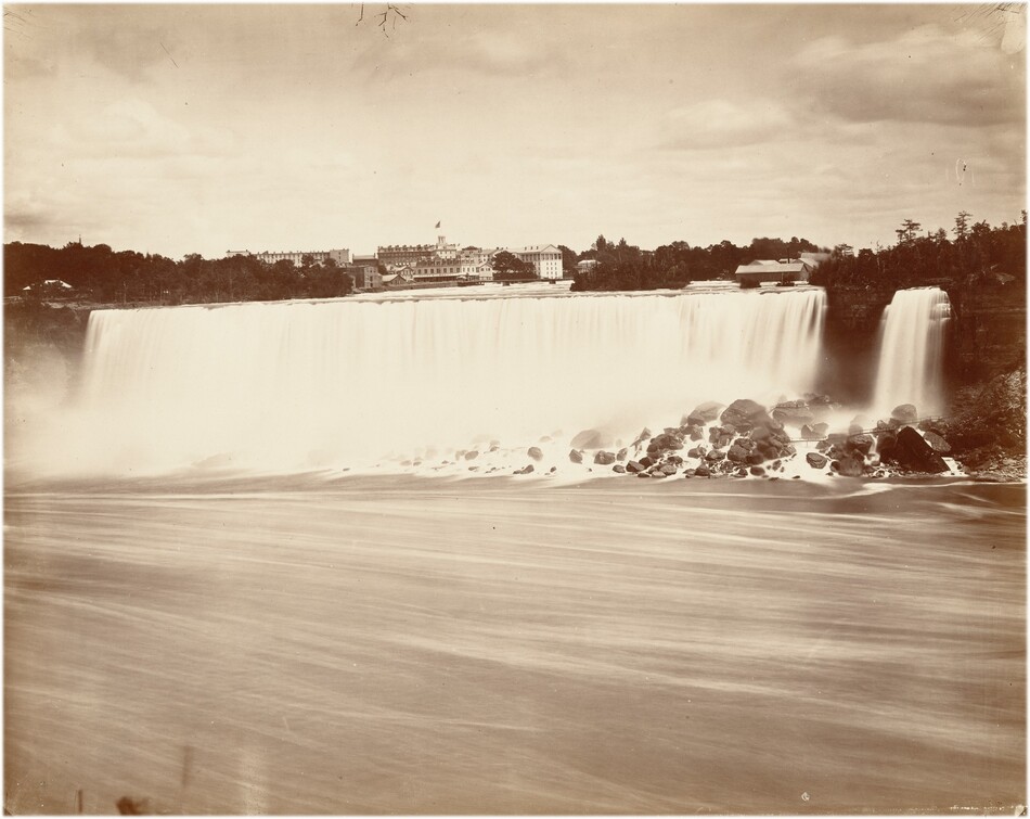 The image shows a sepia-toned view of a large waterfall cascading over a rocky cliff. The horizon is visible at the top with buildings, including one with a flag, silhouetted against the sky. The flowing water appears smooth and misty due to a long exposure, descending from the top of the falls to the river below. Rocks disrupt the water's flow, creating white splashes. The image has a soft, blurred quality due to motion in the water. The color palette is muted and vintage, emphasizing textures and contrasts rather than distinct colors. In the distance, dense trees flank the structures.