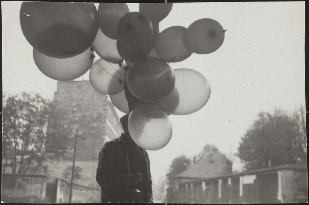 The image shows a person from the head down to the waist. The person is standing and is partially obscured by a bunch of balloons. The face of the person is hidden by the balloons. They are wearing a dark-colored jacket. There are no visible accessories or jewelry, and their hands are partially visible, likely holding the strings of the balloons. The background features a blurry urban setting with old buildings and trees on either side. The image is in black and white.