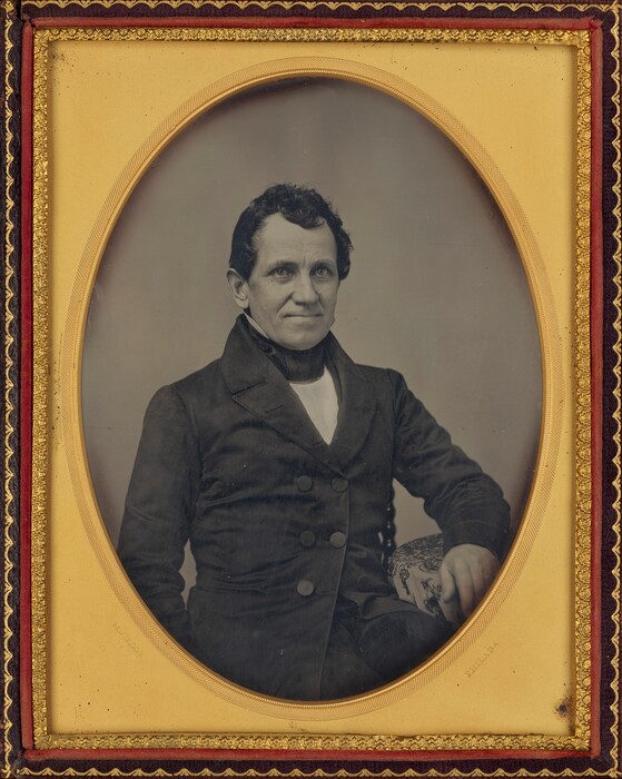 The image shows a man's upper body, from just below the waist upwards. He is slightly turned to his right, sitting with his left arm resting on a surface. The man has short, wavy dark hair and a prominent facial structure with high cheekbones and a slight smile. He wears a dark, double-breasted coat with large buttons over a dark collar and a visible white shirt underneath. The background is plain and neutral. The photograph is bordered by an ornate frame with gold accents.
