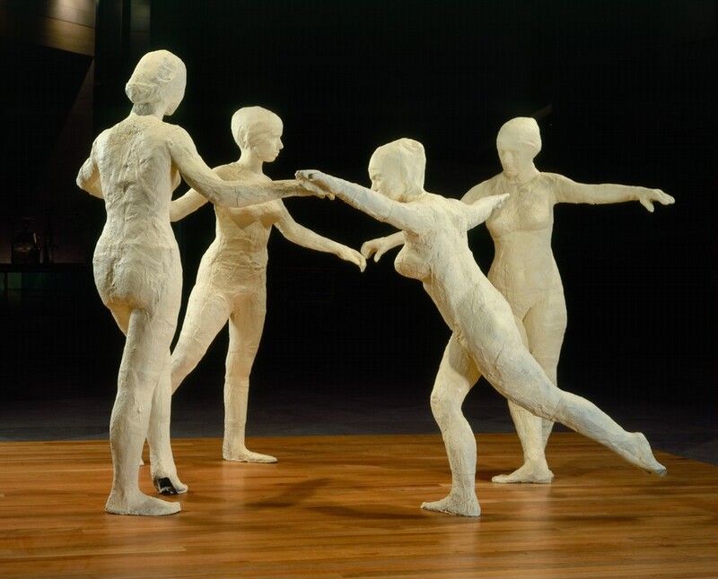 Four ivory-white, free-standing, nude women dance in a circle on a wooden surface against a deeply shadowed background. In this photograph, the two women on the far side of the circle face us, one to our left turns away from us, and the one closest to us faces our left in profile. The one facing our left leans forward with her arms spread wide at shoulder height. Her near leg extends back, and the other leg is bent slightly at the knee. The woman to our left has her back to us. Her right arm reaches out to her right, and she steps forward onto the ball of her left foot. The third woman stands beyond and between the first two. She is angled to our right with her arms spread outward and her right leg, on our left, stepping back. The final woman is on the right and faces us with her head bowed down and to our left. She spreads her arms out just below shoulder-height, and her feet are turned in opposite directions.