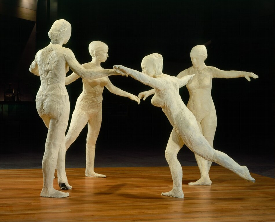 Four ivory-white, free-standing, nude women dance in a circle on a wooden surface against a deeply shadowed background. In this photograph, the two women on the far side of the circle face us, one to our left turns away from us, and the one closest to us faces our left in profile. The one facing our left leans forward with her arms spread wide at shoulder height. Her near leg extends back, and the other leg is bent slightly at the knee. The woman to our left has her back to us. Her right arm reaches out to her right, and she steps forward onto the ball of her left foot. The third woman stands beyond and between the first two. She is angled to our right with her arms spread outward and her right leg, on our left, stepping back. The final woman is on the right and faces us with her head bowed down and to our left. She spreads her arms out just below shoulder-height, and her feet are turned in opposite directions.