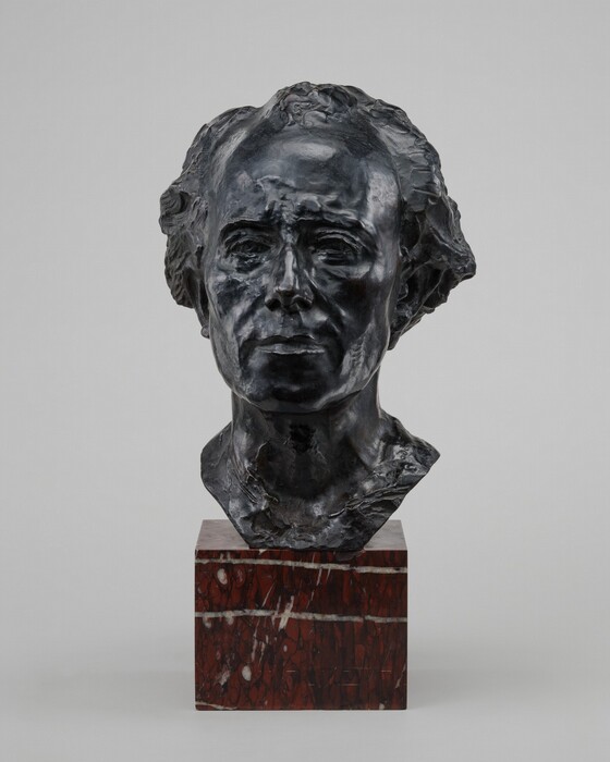 Cast in bronze, this freestanding sculpture shows the head and neck of a man with wavy hair and a deeply lined face. His thick hair appears brushed back from a high forehead. His brows are gathered, his cheeks hollow, and he has a square jawline. The bust stops just below the neck, and the sculpture sits on a maroon-red stone base streaked with white and black.