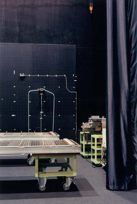 This is a photograph of a room with technical equipment. The room has a dark wall and various technical and industrial equipment. In the foreground, there is a large green cart on wheels with a metallic structure on top. To the right, there is another similar cart with machinery or components. The wall behind the carts has a black pegboard system with pegs and cables. An orange hoist is visible at the top of the frame. The room has an industrial aesthetic, with elements ready for use or in operation.