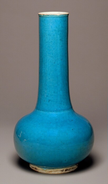 This is a tall ceramic vase against a plain, neutral background. The ceramic has a vivid blue glaze with some slight variations in tone. The vase has a bulbous base and slender neck, with a slightly flared white rim.