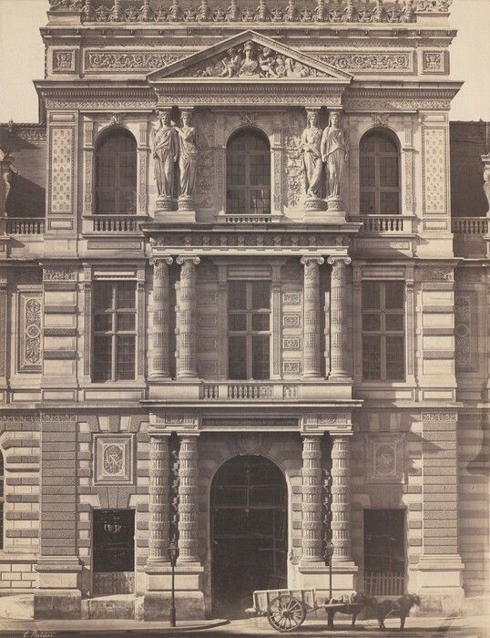 This image shows an ornate and classical architectural facade. It features a grand classical building with intricate detailing, grand columns, a large arched entranceway framed by textured columns, a richly detailed frieze, and statues of robed figures between arched windows. The facade is adorned with ornate sculptures and relief work, including a pediment with carved figures. At street level, a horse-drawn cart is visible.