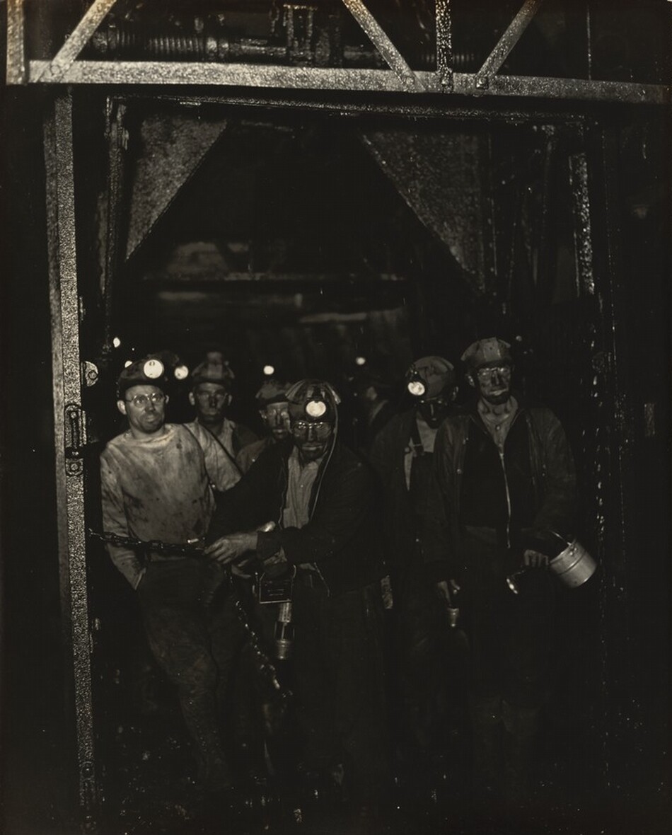 The image shows a group of men standing in a tunnel or mine shaft. The upper half of their bodies is visible, with several standing in the foreground and others in the background. The men are wearing headlamps on their helmets, illuminating their soot-covered faces. Some of the men have short-cropped hair visible beneath their helmets. They are dressed in rugged, dust-stained work clothes suitable for mining, such as overalls and jackets. One man in the front holds a mining drill, while another holds a tin canister. A metal structure is visible in the background, indicating a mining operation setting, with dark and industrial surroundings.