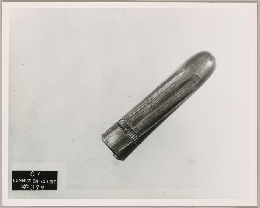 This is a black-and-white photograph of a bullet. The image shows a single bullet against a plain background, tilted diagonally, with the rounded end pointed towards the upper right corner and the flat end pointed to the bottom left corner. The bullet has engraved or grooved lines on its casing and reflects the light. A black label on the bottom left corner reads "C1 COMMISSION EXHIBIT #399". The background is light gray.