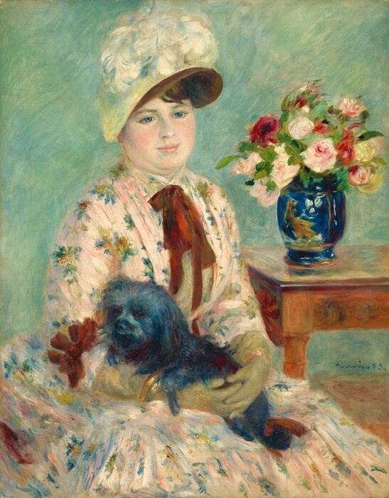 Shown from the lap up, a pale-skinned woman wearing a floral-print dress holds a black lap dog in this vertical portrait painting. The woman’s body is angled to our right, and she gently smiles as she looks slightly down in that direction with blue eyes. She has a round face, rosy, smooth cheeks, pale pink lips, and a double chin. Her brown hair is mostly covered by a hat with a brown brim and pale yellow crown. A bloom of white feathers nestles at the top front of the hat. Her dress is pale pink with a floral pattern painted with dabs of yellow, red, blue, and green. A burgundy-red bow is tied around her neck and lies limply down her chest. The woman wears gold bracelets over pale olive-green gloves that stretch almost to her elbows. Her fingers are interlaced as she holds the shaggy black dog in her lap. The dog’s front paws rest over the woman’s right forearm, to our left. A table in the background to the right holds a wide-mouthed, lapis-blue vase painted with a yellow and orange bird and white flowers. The vase holds at least a dozen pink, red, and white flowers and greenery. The background is painted with strokes of turquoise and mint green. Some brushstrokes are visible, as in the flowers and the woman’s dress, and some brushstrokes are blended, as in the woman’s face. The artist signed the painting near the bottom right corner, “Renoir 83.”