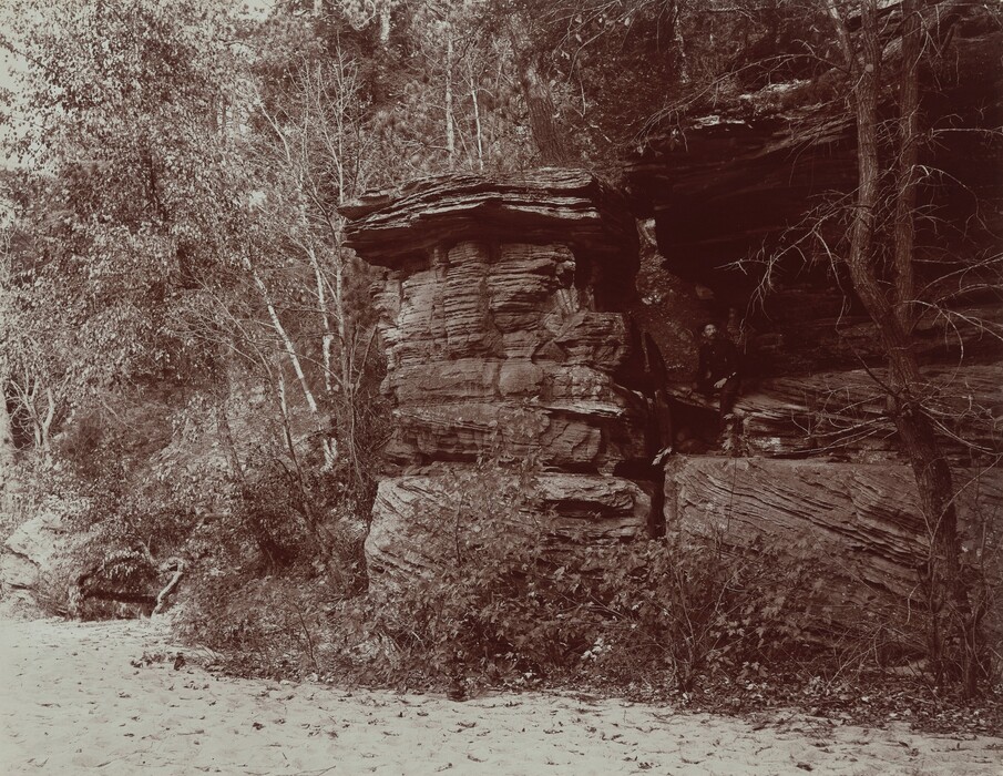 A person sits in a rocky formation at the edge of a dense forest in this horizontal black and white photograph. The rocks are blocky and horizontally or diagonally striated. The person sitting inside has pale skin, a dark mustache, and wears a dark suit. A stretch of sand separates us from the structure.