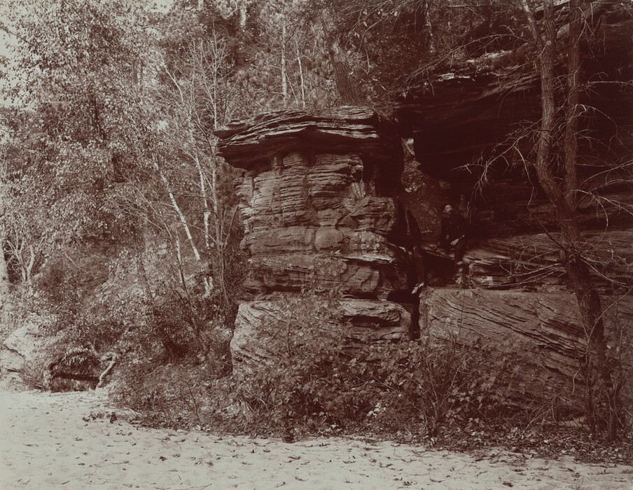 A person sits in a rocky formation at the edge of a dense forest in this horizontal black and white photograph. The rocks are blocky and horizontally or diagonally striated. The person sitting inside has pale skin, a dark mustache, and wears a dark suit. A stretch of sand separates us from the structure.