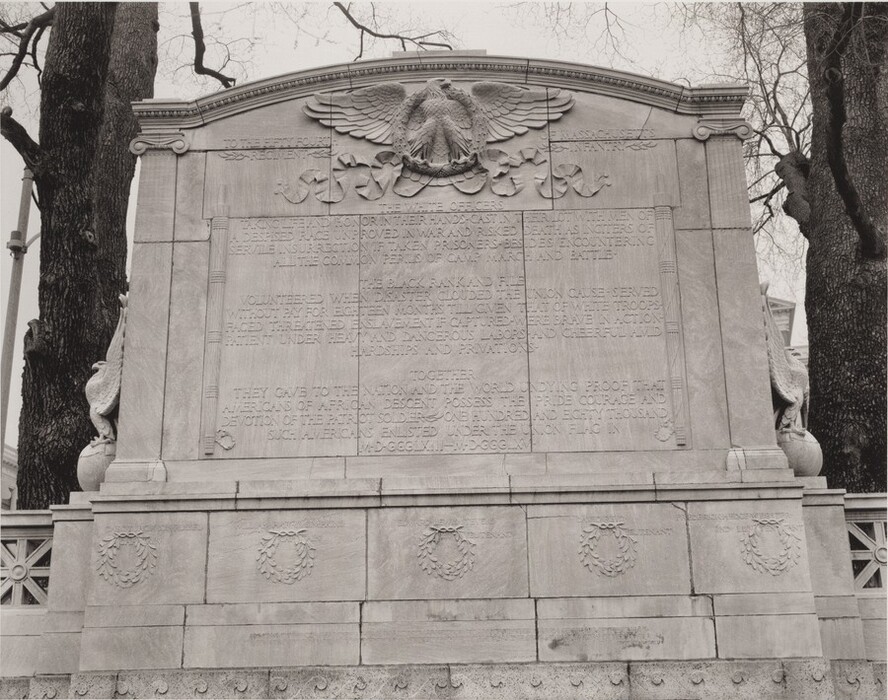 The sculpture is a large, rectangular monument with inscriptions and decorative carvings. The monument likely commemorates the Massachusetts 54th Regiment Infantry. At the top, there is a sculptural relief of an eagle with spread wings. Mythological gryphons are on either side, appearing vigilant and protective. The material is stone, a light color common in historical monuments. It features classical architectural details and is located outdoors in a landscaped area with surrounding trees.
