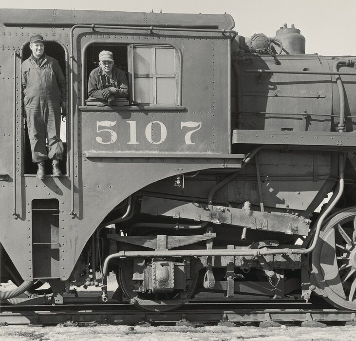 This is a photograph of a steam locomotive engine. The image shows a detailed side view of the train engine, featuring the number "5107" above the wheels. The mechanical details like pistons, pipes, and metal plates are visible, showcasing the engineering of the locomotive. The photo has a classic black-and-white aesthetic.