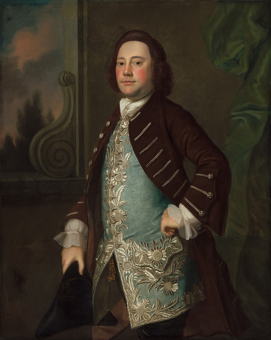 Shown from the knees up, a pale-skinned man with rosy cheeks looks at us in front of a window opening in this vertical portrait painting. The man’s body is angled to our left, but he turns his head to look at us with blue eyes under smooth brows. His chestnut-brown hair is brushed off his high forehead, which slopes down to a wide nose, notably flushed cheeks, and closed pink lips. A white cravat is knotted at his throat, and his left hand, to our right, rests on his hip so it pulls back the dark brown coat he wears. The coat has flaring sleeves and silver embroidery. Translucent ivory-white cuffs of his shirt rest along the backs of his hands. His long aqua-blue vest has a floral brocade pattern and elaborate embroidery of flowers and leaves down the buttoned opening of the front, around the scalloped pocket we see, and along the bottom hem, which comes to about mid-thigh. The man holds a black tricorner hat in his right hand, to our left, by that hip. Shimmering fern-green fabric hangs in folds and swags along the left edge of the painting in front of an ash-brown wall. The rectangular window opening is to our left.  A volute climbs up the lower right corner of the opening, its scrolling bulk resembling a snail’s shell. A twilight sky glows over a few treetops in the landscape beyond. The artist signed the painting as if he had inscribed the front of the windowsill. Text reads, “I: Blackburn Pinx.”
