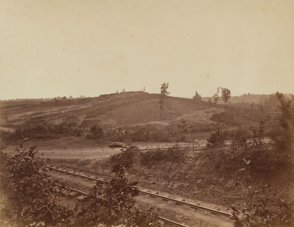This image shows a landscape with railway tracks in the foreground. Beyond the tracks is an area of rough terrain with sparse shrubbery leading to a gently sloping hill in the background. Scattered trees and hints of vegetation are visible, with a man-made structure or fortifications on the horizon. The color tones are predominantly brown and beige, with a pale sky.