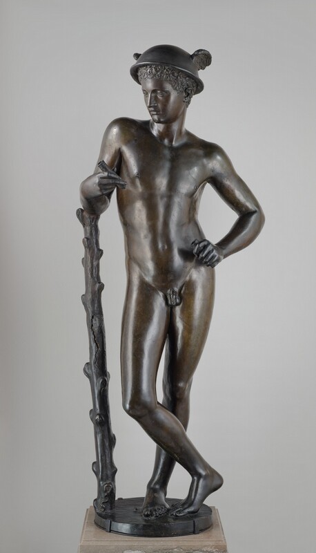 A nude boy or young man standing with one arm leaning on a staff is cast in bronze in this free-standing sculpture. In this photograph, the boy’s body is squared to us, but the head turns to look down to our left. The boy has a straight nose, smooth cheeks, thin lips, and a pointed chin. A cap of tight curls is covered by a bowl-like helmet with wings on either side. The man stands with his weight mostly on his left leg, to our right, which is straight. His other ankle crosses in front of the standing leg, and the toes and ball of that second foot rest lightly on the round base. The man settles into his left hip so his torso sways to our right. His right elbow, to our left, rests on the ribcage-high staff, and his opposite shoulder drops as he rests that hand on his hip. He holds short pipes in both hands. The gentle contours of the boy’s muscles can be made out under his smooth skin. The staff has knobs as if branches had been shorn off. A tiny snake curves in a tight slithering S at the base of the staff, and a couple pebbles are near the boy’s feet. The sculpture rests a square, light brown stone plinth against a pale gray background.
