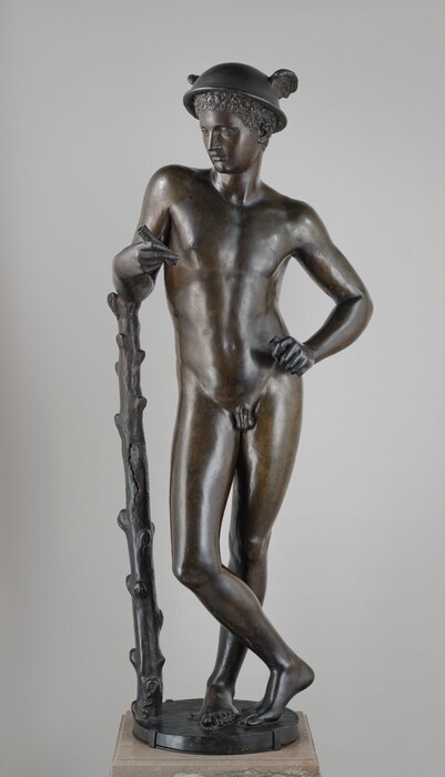A nude boy or young man standing with one arm leaning on a staff is cast in bronze in this free-standing sculpture. In this photograph, the boy’s body is squared to us, but the head turns to look down to our left. The boy has a straight nose, smooth cheeks, thin lips, and a pointed chin. A cap of tight curls is covered by a bowl-like helmet with wings on either side. The man stands with his weight mostly on his left leg, to our right, which is straight. His other ankle crosses in front of the standing leg, and the toes and ball of that second foot rest lightly on the round base. The man settles into his left hip so his torso sways to our right. His right elbow, to our left, rests on the ribcage-high staff, and his opposite shoulder drops as he rests that hand on his hip. He holds short pipes in both hands. The gentle contours of the boy’s muscles can be made out under his smooth skin. The staff has knobs as if branches had been shorn off. A tiny snake curves in a tight slithering S at the base of the staff, and a couple pebbles are near the boy’s feet. The sculpture rests a square, light brown stone plinth against a pale gray background.