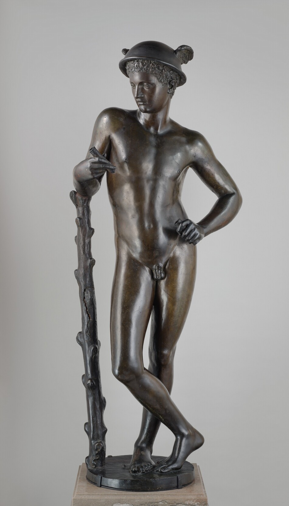 A nude boy or young man standing with one arm leaning on a staff is cast in bronze in this free-standing sculpture. In this photograph, the boy’s body is squared to us, but the head turns to look down to our left. The boy has a straight nose, smooth cheeks, thin lips, and a pointed chin. A cap of tight curls is covered by a bowl-like helmet with wings on either side. The man stands with his weight mostly on his left leg, to our right, which is straight. His other ankle crosses in front of the standing leg, and the toes and ball of that second foot rest lightly on the round base. The man settles into his left hip so his torso sways to our right. His right elbow, to our left, rests on the ribcage-high staff, and his opposite shoulder drops as he rests that hand on his hip. He holds short pipes in both hands. The gentle contours of the boy’s muscles can be made out under his smooth skin. The staff has knobs as if branches had been shorn off. A tiny snake curves in a tight slithering S at the base of the staff, and a couple pebbles are near the boy’s feet. The sculpture rests a square, light brown stone plinth against a pale gray background.