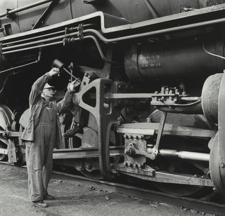 This is a photograph of a steam locomotive. The image captures a detailed view of the mechanical components of a large, robust steam locomotive. The photograph's focus is on the large wheels, pistons, pipes, and joints that make up the undercarriage of the train. The metallic surfaces are covered with a layer of grime and oil. The photograph uses a black and white palette, emphasizing the rugged and functional design of early industrial machinery. The composition of the photograph draws attention to the complexity and intricacies of the locomotive's mechanics.