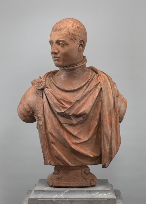 The sculpture depicts a man's bust with short hair, dressed in a draped garment featuring a shell-shaped decoration on his right shoulder. The material resembles terracotta, giving the sculpture a reddish-brown hue. The texture is smooth and detailed, with realistic folds in the clothing and defined facial features. The man's gaze is slightly to his left. The bust is placed on a simple base that complements its style and color. The presentation is highly detailed and lifelike, capturing a sense of presence and historical attire.
