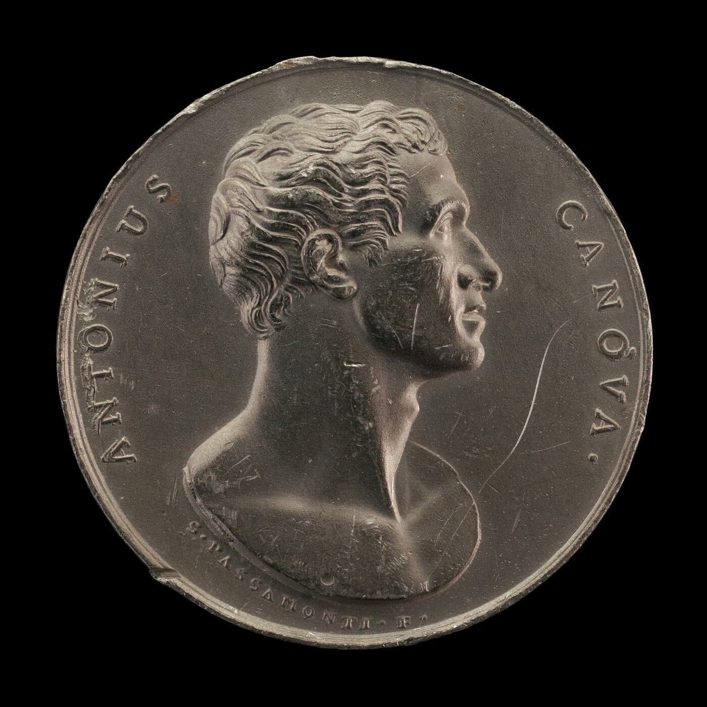 The image shows a portrait sculpture on a circular medallion. The subject is a male figure with textured hair, facing to the right. The sculpture is dark gray and appears to be made of metal. The surface has scratches and realistic texture. Inscriptions on the medallion read "ANTONIUS CANOVA" and "G.PASSAMONTI. F."