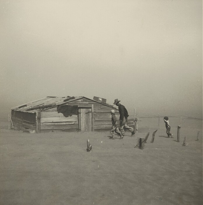 A man and two children dash for a dilapidated, wooden structure in a flat, deserted landscape in this horizontal photograph. The dusty sky and sandy ground blend together in a haze. The man is tall and wears a dark coat and hat. His head tipped down so the brim protects his face, and he leans forward. Next to him is a child wearing a white cap. A few steps behind them, to our right, is another child with his or her hands in the air. The building is a low, A-frame structure made of broken, horizontal wood planks. The gently sloping tin roof is held down by what seems to be larger pieces of wood. The door is in the center of the side we see, and is constructed of vertical planks of wood fastened by dark metal bits. To the left of the door a large hole appears to have been patched in the side of the structure.