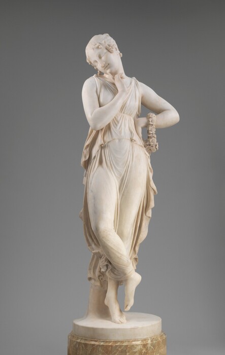 Carved from white marble, a woman balances on the ball of one foot as she holds one finger to her chin in this freestanding sculpture. The woman’s body faces us in this view, but she tips her head down and to our left and she looks in that direction. Ringlets frame her round face, and the corners of her mouth pull back in a slight smile. Her right elbow, to our left, is bent so she can delicately touch her chin with that pointer finger. The back of her other hand is braced against her hip, and a ring of flowers hangs from that forearm. Fabric is gathered over her shoulders, belted under her bust, and falls in delicate folds to her ankles. The woman is barefoot as she lifts one foot and crosses that ankle in front of the standing leg. Perched on the balls of that second foot, the weight of the sculpture is supported by a column behind the woman. She stands on a disk-like, round base on a pedestal made of polished parchment-brown marble. The sculpture is photographed in front of a gray background.