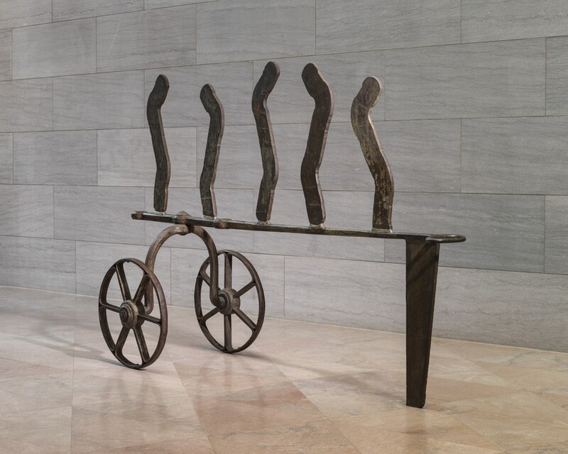 This iron, freestanding sculpture consists of two wheels connected by an upward curving axle, an abstract, carriage-like structure, and five wavy lines standing upright along the top. The wheels each have six spokes and thin rims. In this photograph, the upside-down, U-shaped axle is bolted to the left end of a horizontal strip of iron, which ends to the right with an oval loop. That end of the strip is supported by a tall, narrow, wedge-shaped piece of iron. Five thin iron strips are spaced evenly across the horizontal bar. The top edges are rounded, and they curve in shallow, angular S shapes. The sculpture stands on a pale pink, polished marble floor against a wall with textured gray marble blocks. The scale of the space shows that the sculpture is at least as tall as an average person.