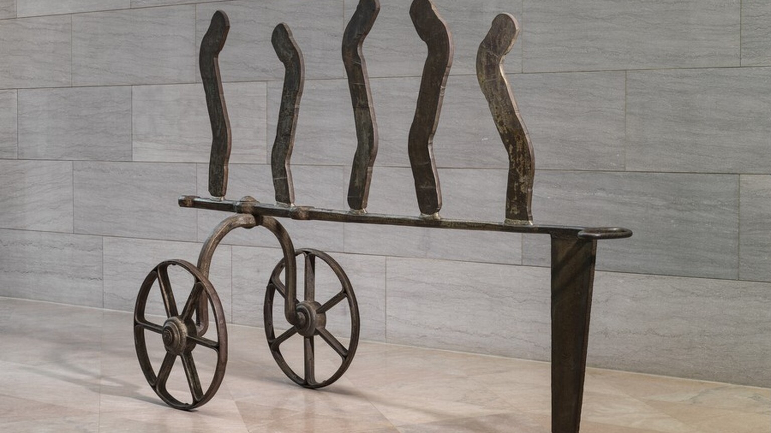 This iron, freestanding sculpture consists of two wheels connected by an upward curving axle, an abstract, carriage-like structure, and five wavy lines standing upright along the top. The wheels each have six spokes and thin rims. In this photograph, the upside-down, U-shaped axle is bolted to the left end of a horizontal strip of iron, which ends to the right with an oval loop. That end of the strip is supported by a tall, narrow, wedge-shaped piece of iron. Five thin iron strips are spaced evenly across the horizontal bar. The top edges are rounded, and they curve in shallow, angular S shapes. The sculpture stands on a pale pink, polished marble floor against a wall with textured gray marble blocks. The scale of the space shows that the sculpture is at least as tall as an average person.