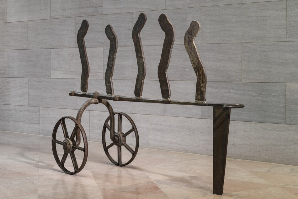 This iron, freestanding sculpture consists of two wheels connected by an upward curving axle, an abstract, carriage-like structure, and five wavy lines standing upright along the top. The wheels each have six spokes and thin rims. In this photograph, the upside-down, U-shaped axle is bolted to the left end of a horizontal strip of iron, which ends to the right with an oval loop. That end of the strip is supported by a tall, narrow, wedge-shaped piece of iron. Five thin iron strips are spaced evenly across the horizontal bar. The top edges are rounded, and they curve in shallow, angular S shapes. The sculpture stands on a pale pink, polished marble floor against a wall with textured gray marble blocks. The scale of the space shows that the sculpture is at least as tall as an average person.