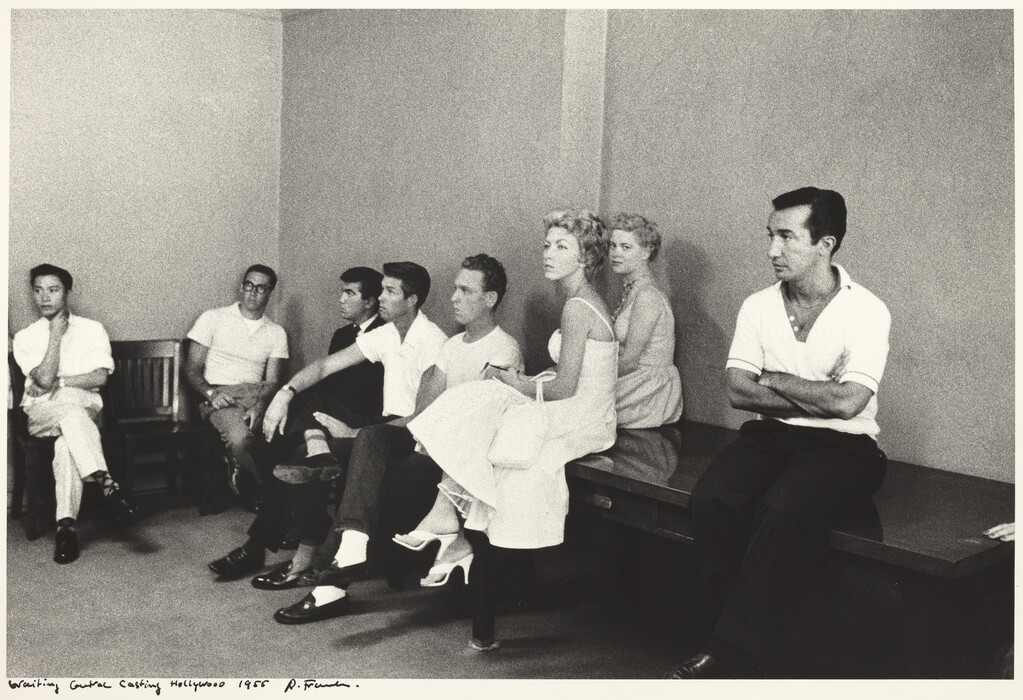 The image shows a group of people sitting in a waiting area. Seven individuals are visible, positioned side-by-side on two benches along the wall. Five men and two women are present. The women, seated on the far right, are wearing light-colored dresses. One woman has short, curly blonde hair, and the other has similar blonde hair. The men wear short-sleeved shirts and dark pants. Two men on either end have their legs crossed. The man on the far left wears a long-sleeve shirt and glasses. The man on the far right has his arms crossed, short dark hair, and a light collared shirt. A man in the middle appears to be holding a cigarette. The room has plain walls and minimal decor.
