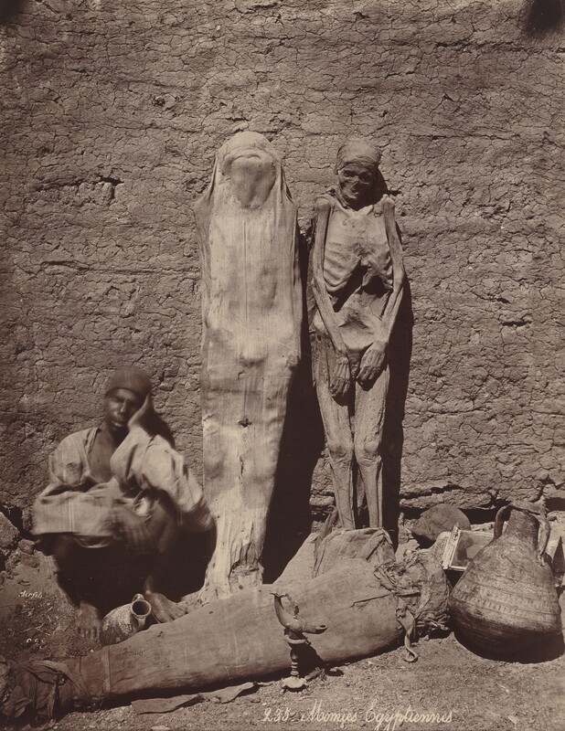 Two wrapped mummies stand propped against a rough, earthen wall in this vertical black and white photograph. The mummy on our left is entirely covered with light-colored fabric while the mummy on the right appears to be uncovered or the coverings have melded to the skin and skeleton. A dark-skinned man sits against the wall to the left of the mummies. He wears a cap and a long, loose, light-colored tunic. His bare feet are planted on the dry, cracked dirt ground, his knees bent so he can prop one elbow on a knee to rest his head, sleeping. His arms and head are slightly blurred, and a vessel sits between his feet. Another mummy, wrapped more thickly than the other two, lies across the bottom of the picture. A larger painted vessel with a wide belly and artifacts, including perhaps a wrapped skull and an oil lamp, are displayed around the prone mummy. Cursive text along the bottom of the photograph reads "235 Momies Egyptiennes."
