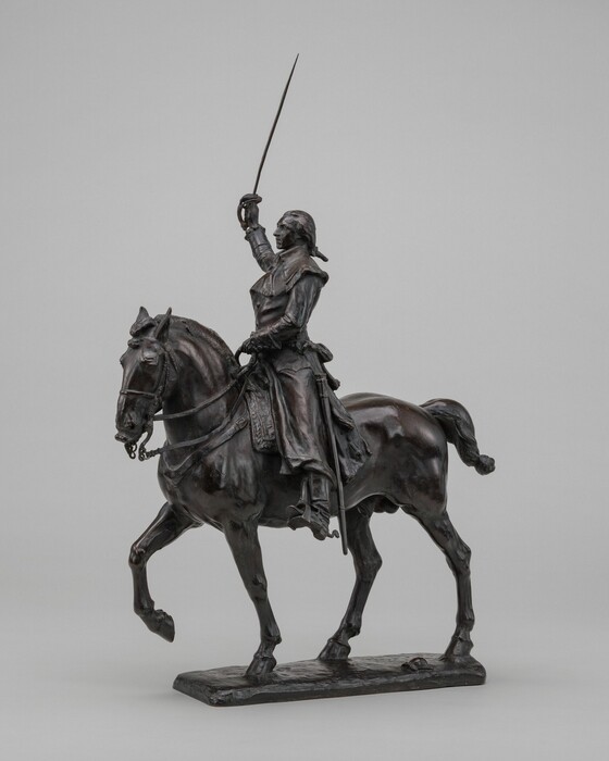 The sculpture depicts a mounted figure of a man riding a horse. The man is dressed in historical military attire and is holding a sword aloft in his right hand. The horse is depicted in motion, with one front leg raised. The sculpture is crafted from a dark material, likely bronze, which emphasizes the intricate details of the rider's and horse's features, such as the flowing mane, the attire on the rider, and the elaborate saddle, against the lighter background.